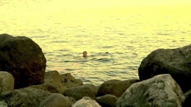 Genç bir kadın günbatımında kayaların arasındaki denizde yüzüyor