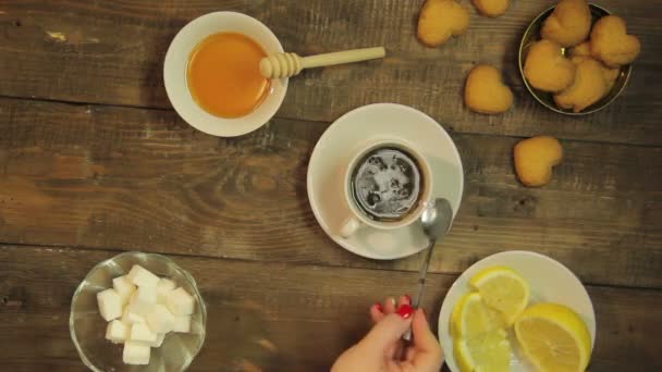 Mano femenina revolviendo con té una cuchara en una taza blanca sobre una mesa de madera — Vídeo de stock