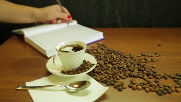 Koppen sterke koffie, een verstrooiing van koffiebonen op de tafel en een vrouw schrijft in een dagboek — Stockvideo