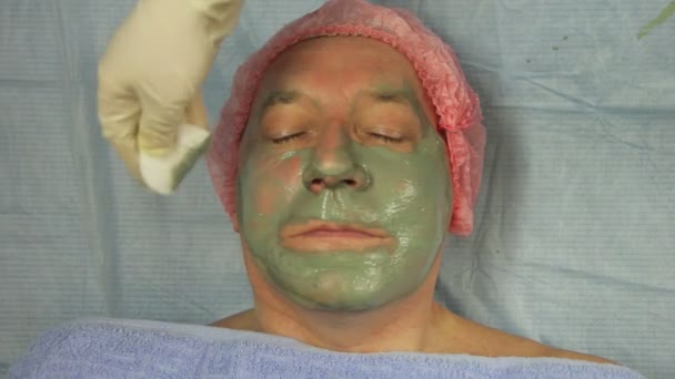 Las manos de un cosmetólogo quitan una máscara de barro de la cara de un hombre con una almohadilla de algodón — Vídeo de stock