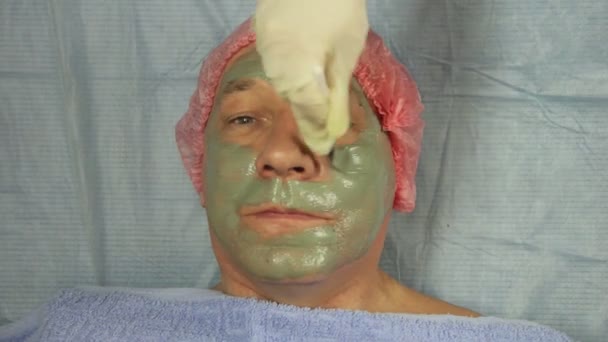 Een vrouwelijke schoonheidsspecialist in handschoenen is de toepassing van een modder masker op het gezicht van een mannelijke clien — Stockvideo