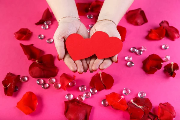 Rode dubbele papier hart in vrouwelijke handen op een roze achtergrond omgeven door rozenblaadjes. — Stockfoto