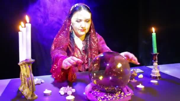 Een zigeunervrouw in een rode jurk in een kamer voor waarzeggerij in soesjes van rook luidt de toekomst in een spiegel bal op tafel — Stockvideo