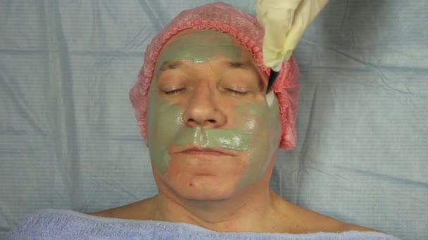 Een vrouwelijke schoonheidsspecialiste in handschoenen zet op een mans een modder masker gezicht — Stockvideo