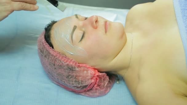 Um esteticista em luvas coloca uma máscara cosmética no rosto de uma mulher. Vista lateral — Vídeo de Stock