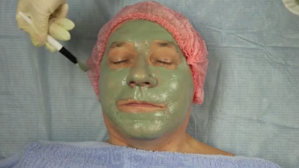 手袋の女性美容師は、男性クライアントの顔にブラシで泥マスクを置きます — ストック動画