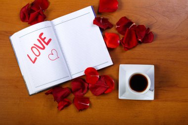 Bir fincan kahve ve bir günlük kağıt ahşap bir masa üzerinde kırmızıyla yazılmış kelime aşk ile gül yaprakları ile kaplı.