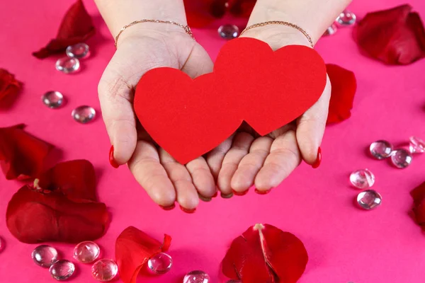 Red double paper heart in female hands on a pink background surrounded by rose petals. — Stock Photo, Image