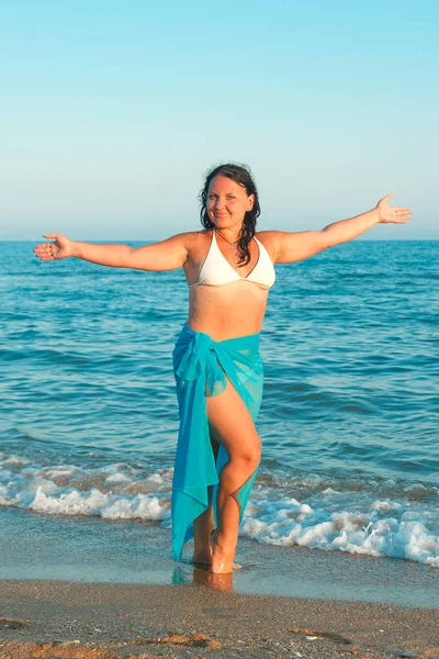 En ung kvinna på stranden mot bakgrund av havet i en vit baddräkt och blå sarong. — Stockfoto