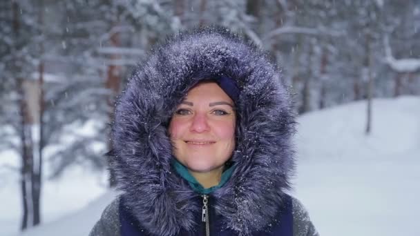 Uma jovem sorri em um capuz em um parque de inverno em uma queda de neve — Vídeo de Stock