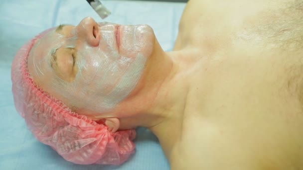 Une femme cosmétologue portant des gants applique un masque aux algues sur le visage d'un homme avec une brosse — Video