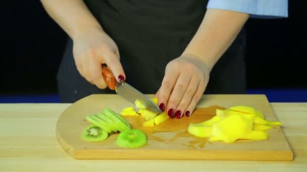 Ženské ruce řezané kousky kiwi a mango na dřevěném prkénku — Stock video