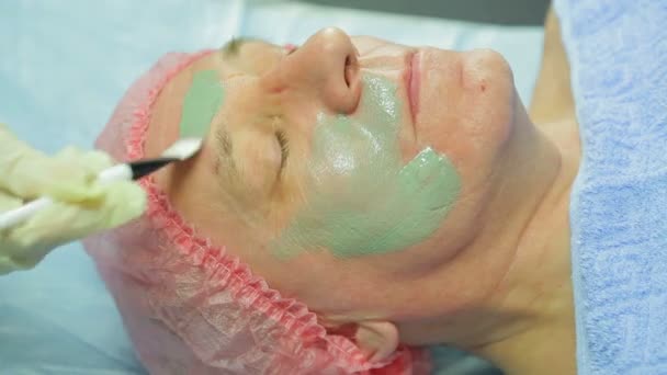 Una cosmetologa donna in guanti applica una maschera di argilla terapeutica al viso di un uomo con un pennello — Video Stock