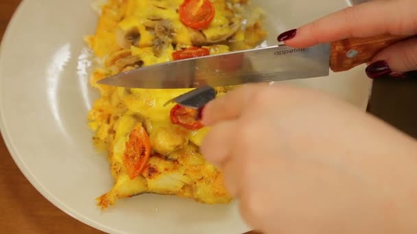 Una mujer con un cuchillo y un tenedor corta queso de pescado al horno con champiñones y tomates cherry en un plato blanco — Vídeo de stock