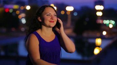 Mavi elbiseli bir genç kadın bir smartphone sokak ışıkların yan bulanık bir arka plan üzerinde konuşuyor. Orta planı. Hareketli kamera.
