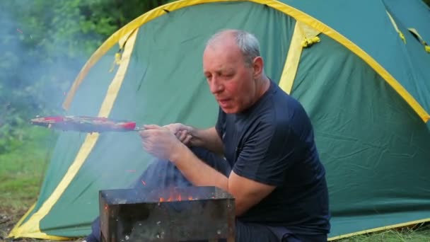 一名身穿蓝色 T恤的男子坐在森林里 坐在一个绿色帐篷的背景下 在火盆里吹着燃烧的煤 中间计划 — 图库视频影像