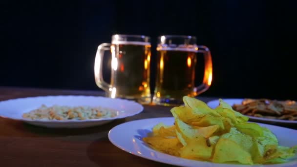 Tasses à bière avec bière en mousse sur fond noir sur une table en bois avec des collations . — Video