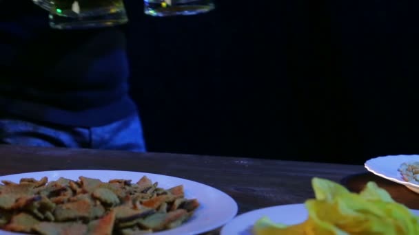 A female bartender puts on a table with snacks on a black background two full mugs of foaming beer — Stock Video