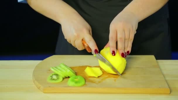 Frauenhände schneiden Kiwi- und Mangostücke auf einem Holzbrett. — Stockvideo