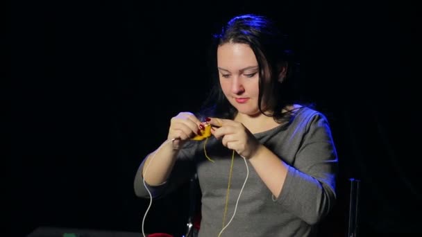 Een jonge brunette vrouw op een zwarte achtergrond breit op rondbreinaalden breien van gele draad — Stockvideo