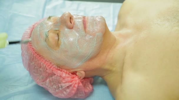 A female cosmetologist wearing gloves applies a seaweed mask to a man s face with a brush. Side view — Stock Video