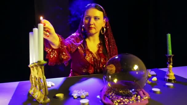 Una mujer gitana con un vestido rojo en una sala de adivinación enciende velas en un candelero tallado — Vídeos de Stock