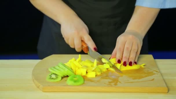 Des mains féminines coupent des morceaux de kiwi et de mangue sur une planche en bois . — Video