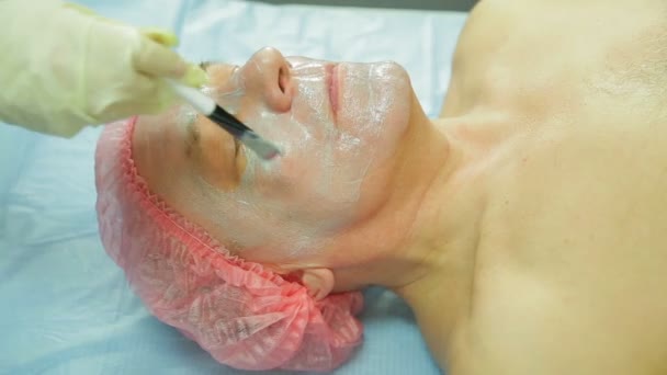 A female cosmetologist wearing gloves applies a seaweed mask to a man s face with a brush. Side view — Stock Video