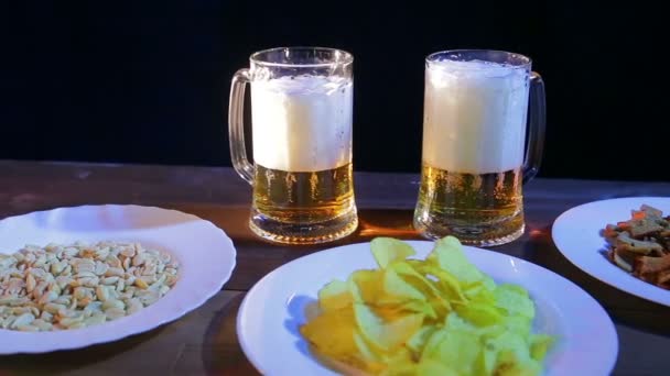 Dos tazas con cerveza espumosa sobre una mesa de madera en un bar sobre un fondo negro. Cerca de la placa con patatas fritas y nueces — Vídeos de Stock