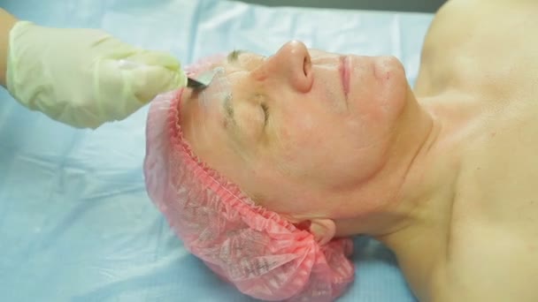 Eine Kosmetikerin in Handschuhen trägt mit einem Pinsel eine Maske auf das Gesicht eines Mannes auf. Seitenansicht — Stockvideo