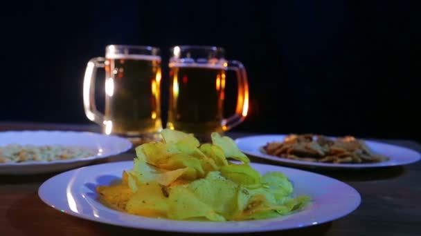 Tazas de cerveza con cerveza de espuma sobre fondo negro sobre una mesa de madera con snacks — Vídeo de stock