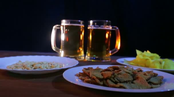 Пивні чашки з пінним пивом на чорному тлі на дерев'яному столі з закусками — стокове відео