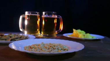 Bira bardağı köpük bira renk ışık aperatifler ile ahşap bir masa üzerinde siyah bir arka plan ile. Ortalama planı