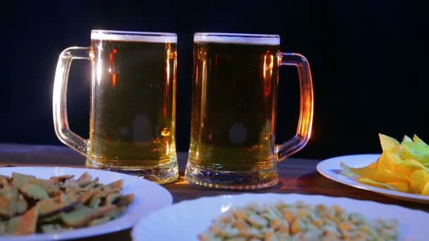 Tazas de cerveza con cerveza de espuma sobre fondo negro sobre una mesa de madera con snacks — Vídeo de stock