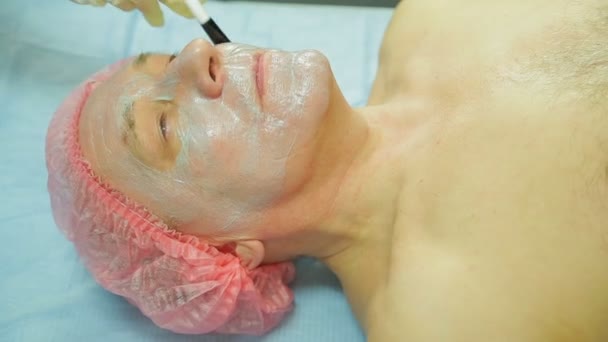 Женщина-косметолог в перчатках наносит маску из водорослей на лицо мужчины кисточкой. Вид сбоку — стоковое видео