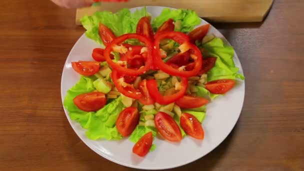Женская рука кладет кольца красного перца в тарелку с салатом и помидорами — стоковое видео