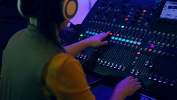 Femme DJ dans un casque derrière une console de mixage travaillant dans l'éclairage couleur. Filmer de l'arrière — Video