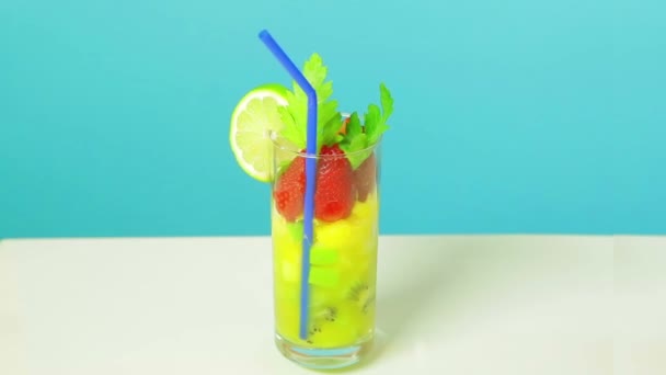 Fruchtcocktail in einem Glas Kiwi, Apfel- und Erdbeerscheiben mit Limette und blauem Strohhalm auf blauem Hintergrund. Glas dreht sich im Kreis. — Stockvideo