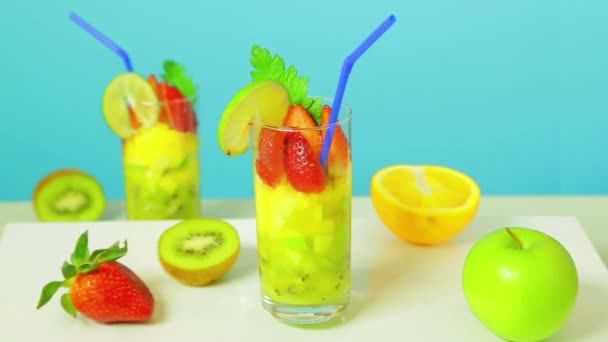 Fruktsallad i glas av kiwi, äpple och jordgubb skivor inredda med kalk med en blå Halm på en blå bakgrund. Glas roterar i en cirkel — Stockvideo