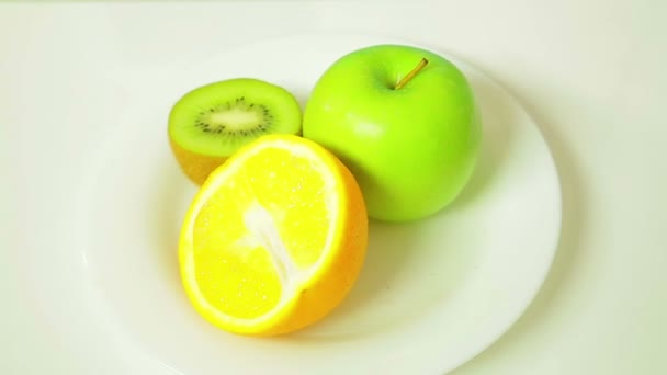 Las mitades de kiwi naranja y manzana verde en un plato blanco giran en círculo — Vídeo de stock
