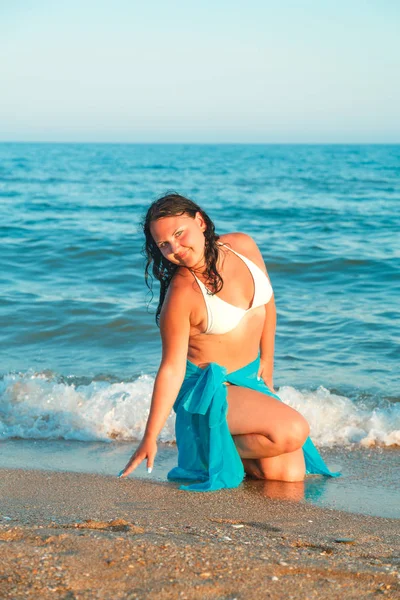 En ung kvinnlig modell sitter på stranden mot bakgrund av havet i en vit baddräkt och en blå sarong. — Stockfoto