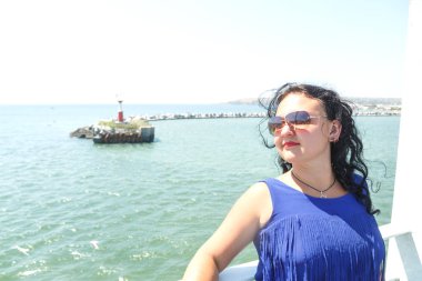 Esmer kadın mavi elbise ve kıyıdan yelken bir motorlu gemi güverte üzerinde güneş gözlüğü.