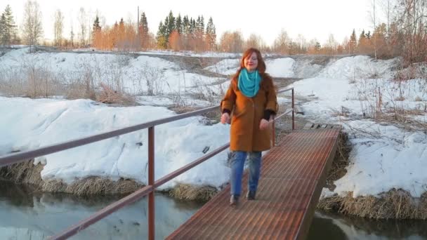 Unga gladlynt rödhårig kvinna korsar en liten flod längs bron i början av våren vid Sol uppgången — Stockvideo