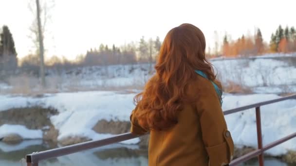 En ung rödhårig kvinna korsar en liten flod längs bron i början av våren vid Sol uppgången. — Stockvideo