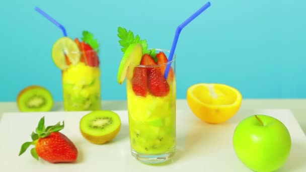 Fruit cocktail in a glass of kiwi, apple and strawberry slices decorated with lime with a blue straw on a blue background. Glass rotates in a circle — Stock Video