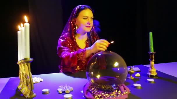 Una mujer gitana con un vestido rojo en una sala de adivinación enciende velas en un candelero tallado . — Vídeo de stock