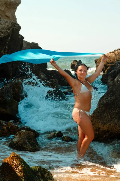 En ung kvinna i en vit baddräkt i havet bland klipporna innehar en blå sarong som flyger över huvudet. — Stockfoto