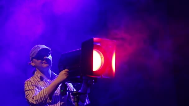 Una joven iluminadora en el escenario trabaja con un proyector de perfil teatral en nubes de humo — Vídeo de stock