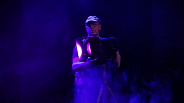 Un hombre en el escenario trabajando con un proyector de perfil teatral dirige la luz en una nube de humo — Vídeo de stock