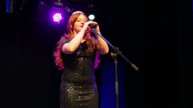 Parlak siyah elbiseli genç kahverengi saçlı aktris, sahnede sahnede bir duman içinde şarkı lar.